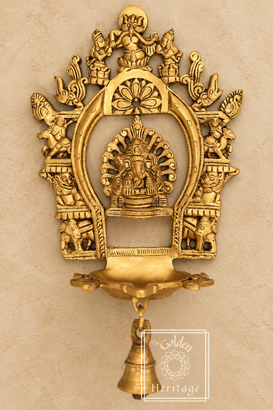 A brass idol of Sukhasana Ganapati with a Prabhavali frame and a hanging Diya (lamp). The idol has multiple arms, each holding different symbols, and is flanked by two goddesses.