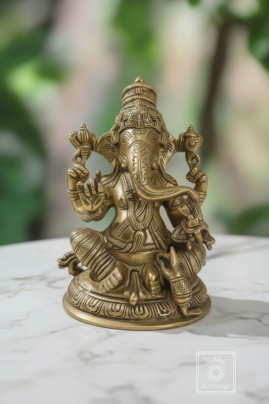 A brass statue of Lord Ganesha, also known as Vinayaka, seated with his right hand held up in the abaya mudra, holding a noose and laddus, with a trident and design carved onto the base of his trunk with his vehicle, the mouse, resting on his left leg.