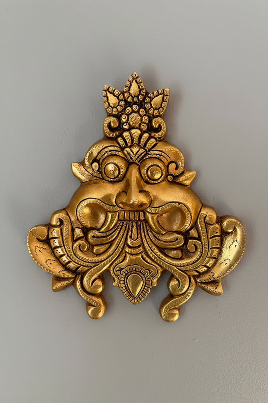 An ornate gold-colored metal wall plaque depicting a stylized face with a crown, photographed against a plain gray background.