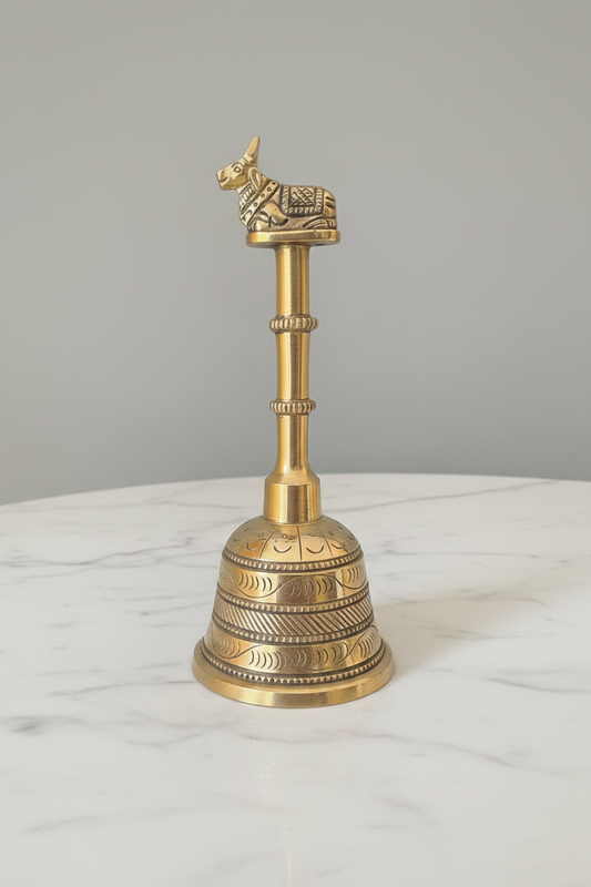 An ornate brass bell with a decorative brass bull figurine on top, displayed on a white marble surface against a neutral gray background.