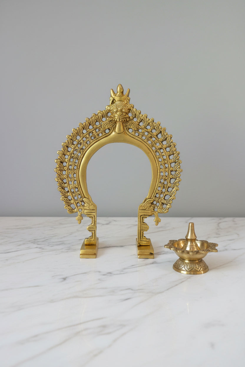 A golden brass arch-shaped decorative sculpture with intricate leaf-like filigree details and a small figure at the top, displayed on a dark reflective surface against a plain gray wall.