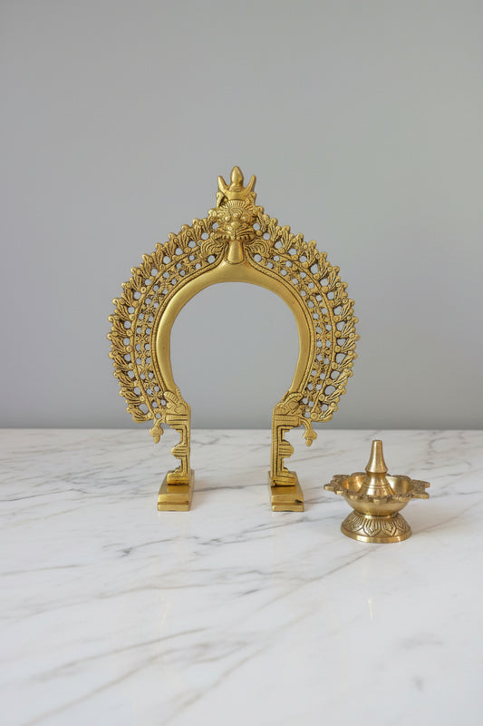 A golden brass arch-shaped decorative sculpture with intricate leaf-like filigree details and a small figure at the top, displayed on a dark reflective surface against a plain gray wall.