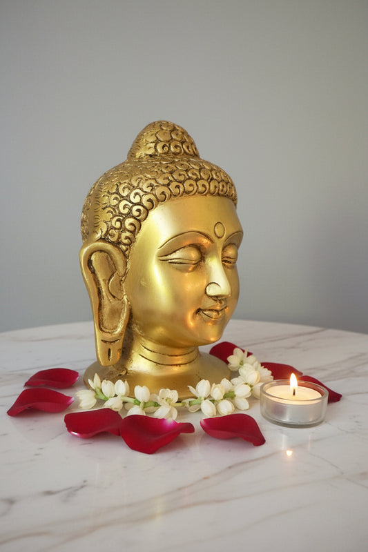 A golden Buddha head bust sculpture with detailed curls in its hair, shown in profile on a dark reflective surface against a plain gray background.