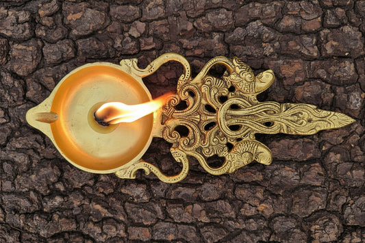 An ornate brass oil lamp with a decorative filigree handle and a small spout, photographed from above on a textured dark brown bark background.
