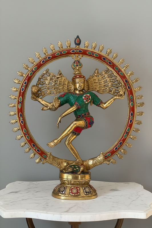 A gold-toned Nataraja statue of Lord Shiva in a dancing pose, adorned with a green shirt, red pants, and a red circular frame with colorful accents, displayed on a white marble-topped table against a gray wall.