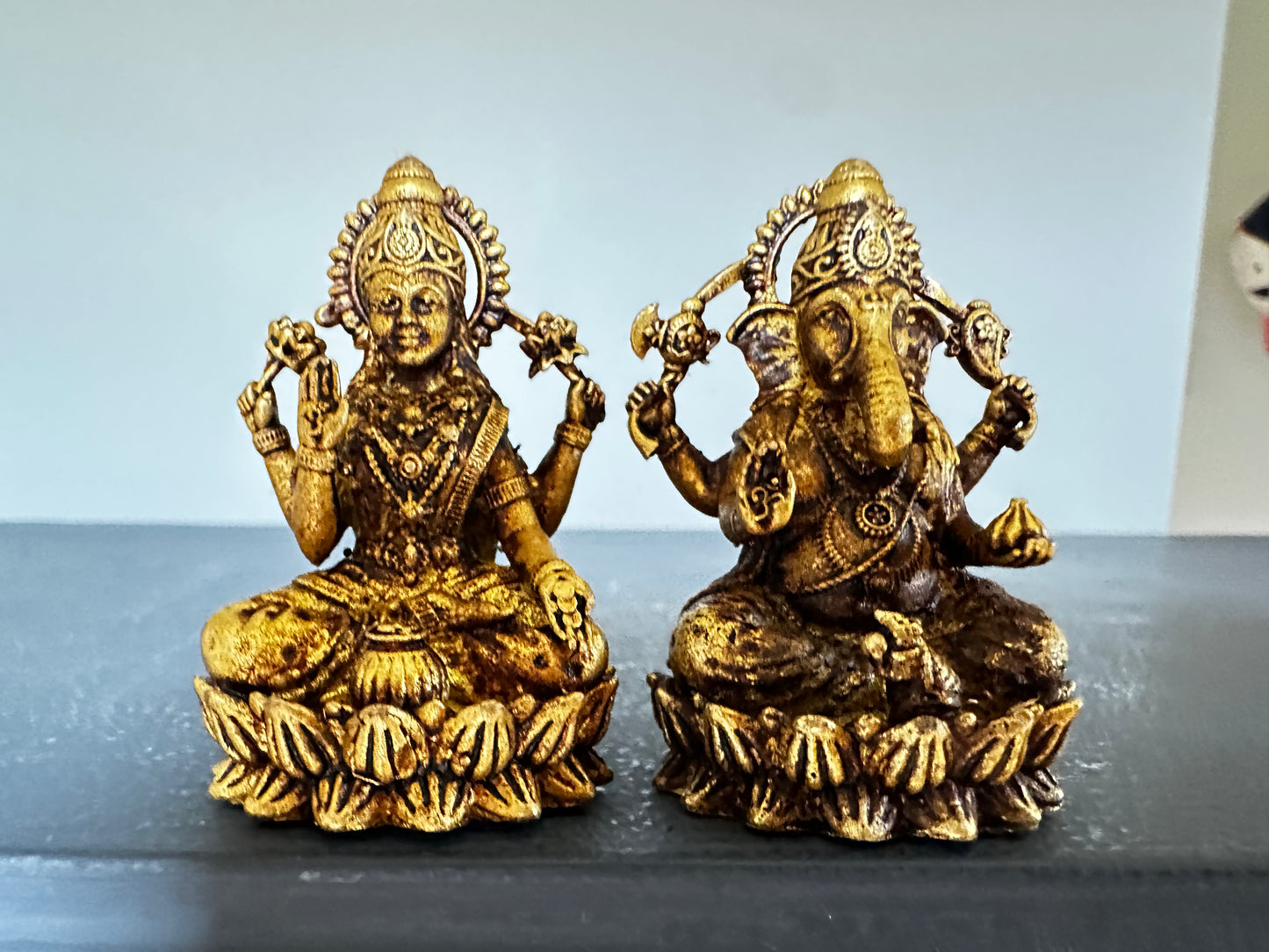 A set of two brass idols, one representing Ganesha and the other representing Lakshmi, both with ornate detailing and seated cross-legged on a platform.