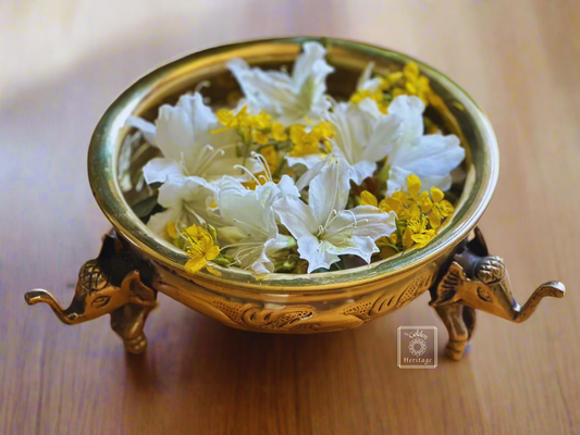A brass decorative bowl with elephant legs design, containing water and flower petals, placed on a wooden surface.