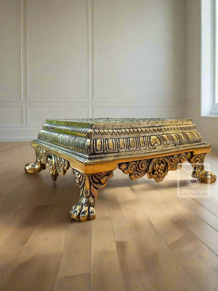 A brass pooja chowki with intricate carvings and a golden finish, placed on a wooden surface.