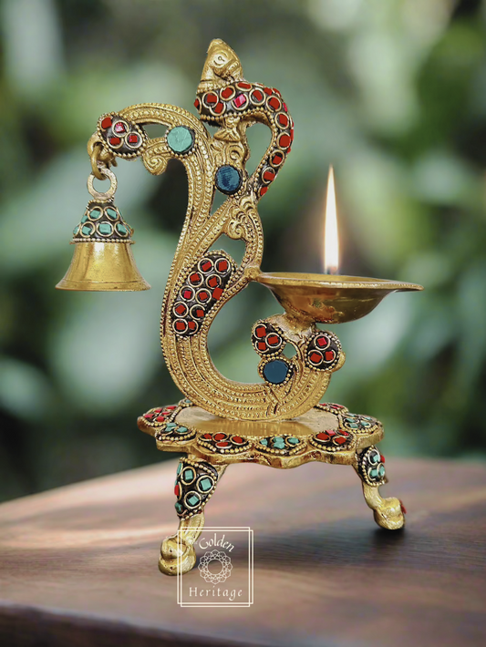 A brass pooja lamp with a peacock design and stone work, featuring a lit candle, bell, and intricate patterns.