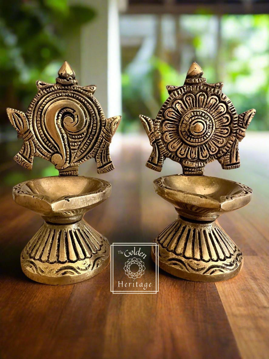 A set of two brass lamps with intricate designs, featuring symbols resembling a conch shell and a chakra wheel, in a gold finish 