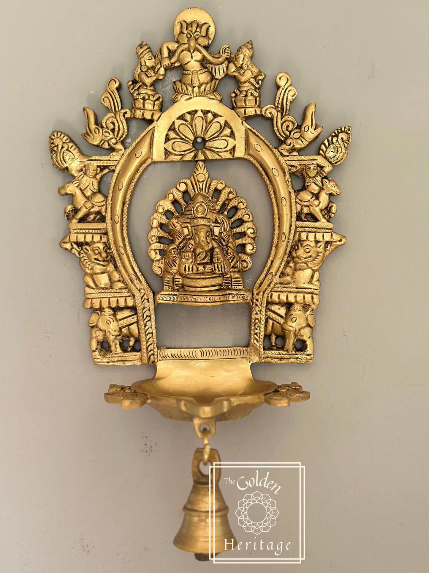 A brass idol of Sukhasana Ganapati with a Prabhavali frame and a hanging Diya (lamp). The idol has multiple arms, each holding different symbols, and is flanked by two goddesses.