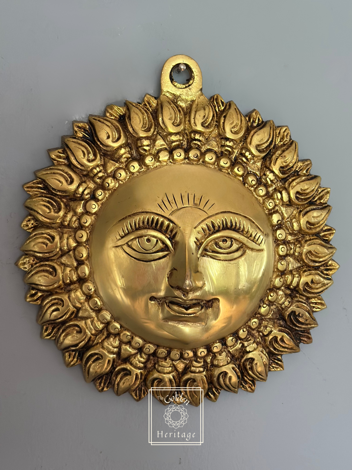 A metallic gold-colored wall hanging depicting the face of Lord Surya with multiple rays around it.