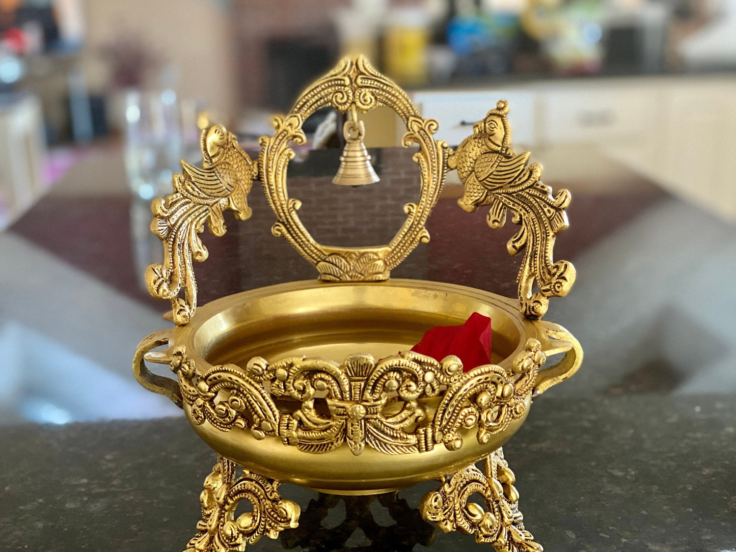A gold-colored metal urli bowl with peacock design and a bell, decorated with flower petals.