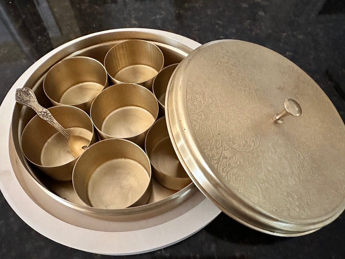 A traditional Indian brass spice box with multiple compartments and a lid, placed on a white surface with a spoon inside one of the compartments.